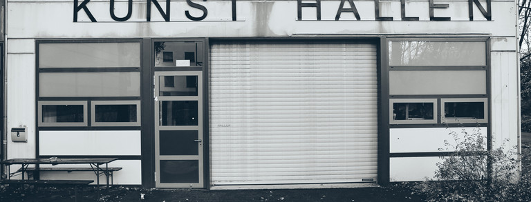 Zu sehen ist der Eingangsbereich der Kunst Hallen von Außen. Über dem Rolltor steht mit schwarzen Holz-Lettern "Kunst Hallen"