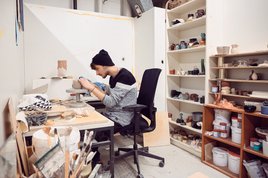 Zu sehen ist der Student Yannick Wetzel in seinem Atelier. Er arbeitet an einer Tonskulptur. Im Hintergrund sind weitere Keramikarbeiten zu sehen. 