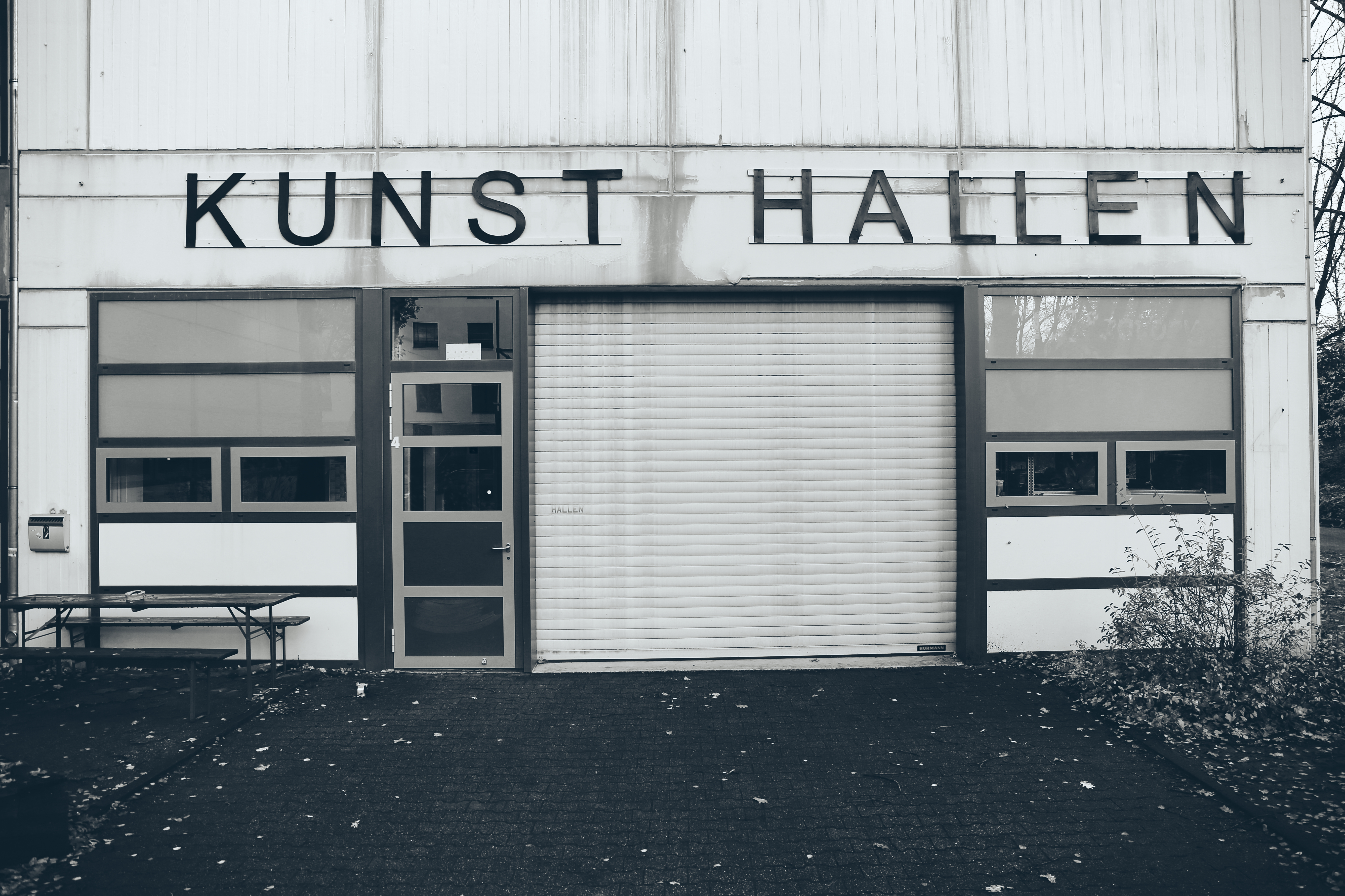 Zu sehen ist der Eingangsbereich der Kunst Hallen von Außen. Über dem Rolltor steht mit schwarzen Holz-Lettern "Kunst Hallen"